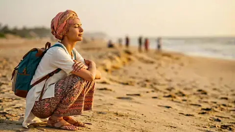 Woman in a patterned headscarf sitting on a beach at golden hour, eyes closed, peaceful smile